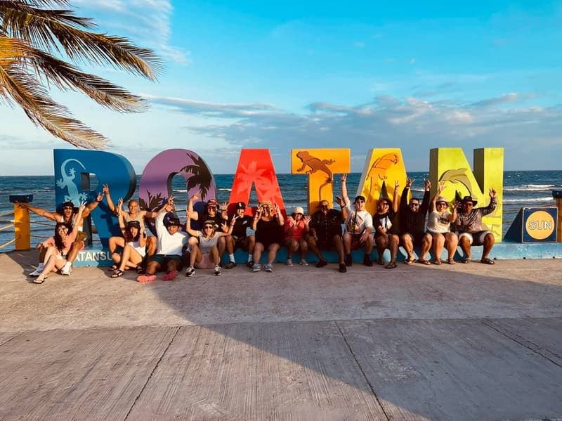 About twenty guests sitting along the giant coloured ROATÁN letters by the sea at sunset, arms raised